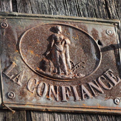 Rusty metal plaque mounted on weathered wood, featuring an oval relief of a man with a dog and the text 'LA CONFIANCE'.