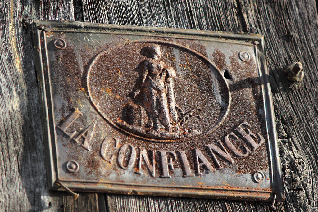 Rusty metal plaque mounted on weathered wood, featuring an oval relief of a man with a dog and the text 'LA CONFIANCE'.